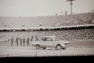 Vintage black-and-white shot of a stadium event featuring a white sedan with "7010" and "Bulgaria" decals driving onto a fiel...