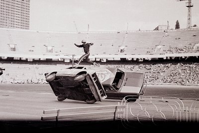 Stunt performer mid-air flips vintage car atop inverted truck in stadium setting, likely 1960s-70s. Olympic rings logo visibl...