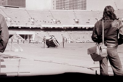 Empty stadium under construction, featuring tiered concrete seating and exposed scaffolding. Two figures in mid-20th-century ...