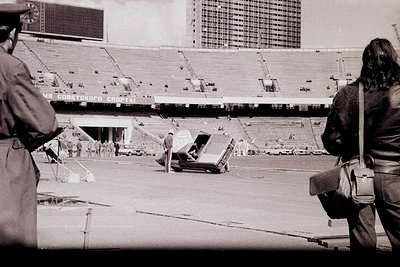 Vintage black-and-white photo of a large stadium under construction, featuring exposed concrete seating and skeletal structur...