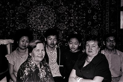 Five adults pose indoors against floral-patterned wallpaper, likely 1970s–1980s. Two women in floral blouses, a man in a chec...
