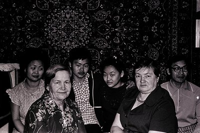 Five individuals pose indoors against a wall adorned with intricate, symmetrical embroidered patterns. The setting appears to...