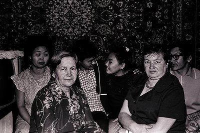 Black-and-white portrait of five adults in a dimly lit interior, likely mid-20th century. Central figure wears floral-pattern...
