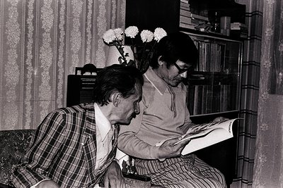 Two men engrossed in reading in a vintage indoor setting, likely mid-20th century. Floral arrangement on a side table, patter...
