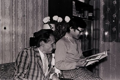 Two men engrossed in reading indoors, likely mid-20th century. Patterned wallpaper and floral vases with white flowers frame ...