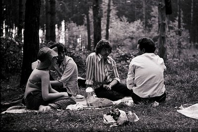 Mid-20th century picnic in a dense forest setting. Four individuals seated on a blanket, surrounded by tall trees and natural...