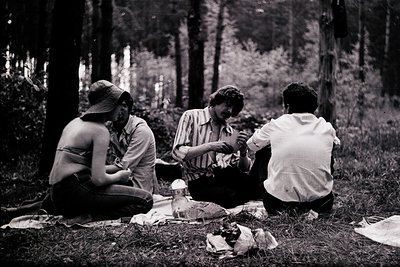 Black-and-white snapshot of three individuals picnicking in a wooded area, mid-20th century. Lush greenery surrounds a blanke...