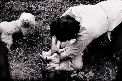A candid black-and-white shot of a person and small white dog in an outdoor setting, likely mid-20th century. The individual,...