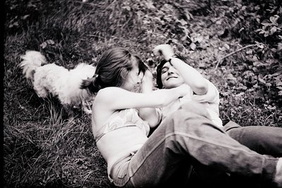 Black-and-white candid shot of two people reclining in grassy field, one shirtless, embracing. Fluffy dog lies nearby. Soft f...