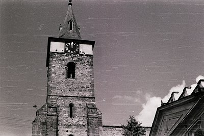 Historic stone church tower with clock and bell niche, framed by power lines. Architectural details include arched windows an...