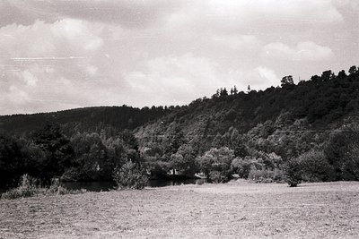 Rural landscape featuring a serene pond surrounded by dense forest on a hillside. Mid-20th century black-and-white photo capt...