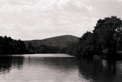 Vintage black-and-white lakeside scene with calm water reflecting forested hills. A lone figure stands near the shore, surrou...