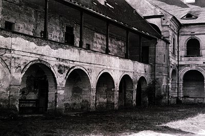 A weathered courtyard arcade with arched openings, likely part of a historic European monastery or cloister. Stonework shows ...