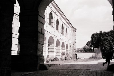 Historic stone archway framing a courtyard with arched colonnade, likely part of a monastery or institutional complex. Archit...