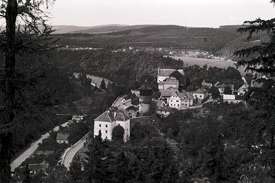 Historic hilltop village with clustered stone buildings and winding road, framed by dense forest. Black-and-white aerial pers...