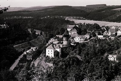 Aerial view of a historic village nestled in a forested valley, likely Eastern Europe. Central structure appears to be a fort...