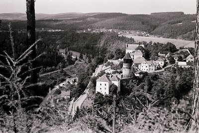 Mid-20th century black-and-white aerial view of a European village nestled in hilly terrain. Prominent stone castle with coni...