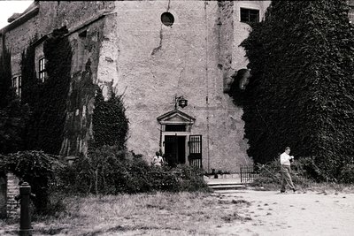 A weathered, two-story stone building with crumbling plaster and overgrown ivy, featuring a small classical-style entrance do...