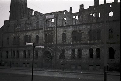 Dilapidated multi-story brick building with Gothic Revival arches, partial roof collapse, and exposed interior framework. Str...
