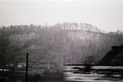 Black-and-white landscape shot from a moving train, showcasing a misty hillside with sparse winter trees and a lone house bel...