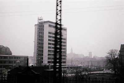 High-angle black-and-white shot of a mid-20th-century urban skyline. Dominated by a Brutalist-style high-rise under construct...
