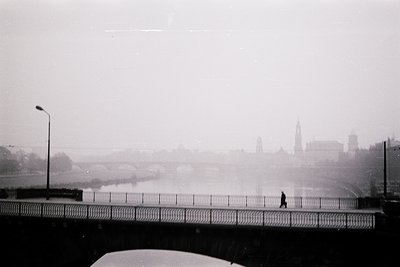 Black-and-white urban scene featuring a lone pedestrian crossing a bridge over a foggy river. Distant cityscape with spires a...