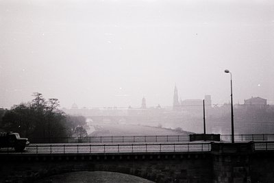 Vintage black-and-white shot of a foggy urban bridge with a single streetlamp, leading toward a distant cityscape. Distinctiv...