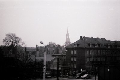 Vintage black-and-white street scene featuring early 20th-century European architecture. Prominent multi-story brick building...