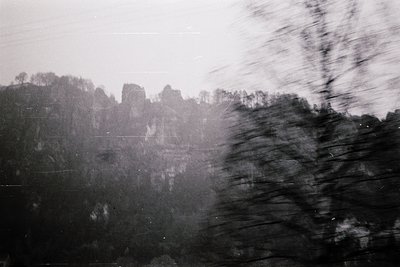 Blurred monochrome shot of a misty, forested hillside with faint ruins visible through fog. Motion blur suggests rapid moveme...