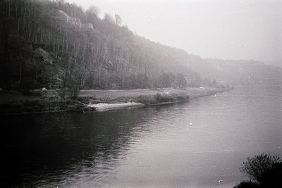 Vintage black-and-white shot of a serene river winding through dense forest. Mist rises from water, partially obscuring rocky...