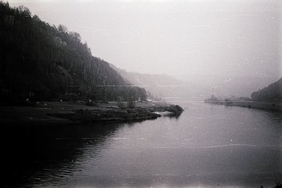 Vintage black-and-white river scene with misty, overcast skies. Dense forest lines both banks, framing a calm, reflective wat...
