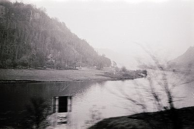 Black-and-white shot of a misty lakeside scene with forested hills in mid-ground. Reflections blur on water’s surface, sugges...