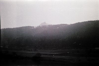 Vintage black-and-white landscape featuring misty, snow-capped mountain range with dense forest below. Fog obscures mid-groun...