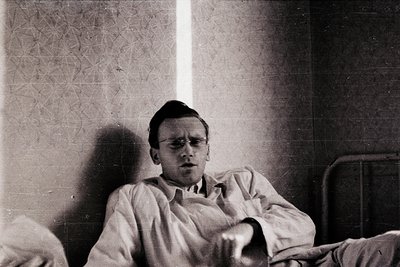 Vintage sepia-toned portrait of a young man in a hospital gown, seated on a metal-framed bed. Geometric wallpaper and a windo...
