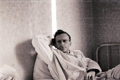 Portrait of a man in a hospital gown leaning against a tiled wall, likely mid-20th century. The clinical setting and vintage ...