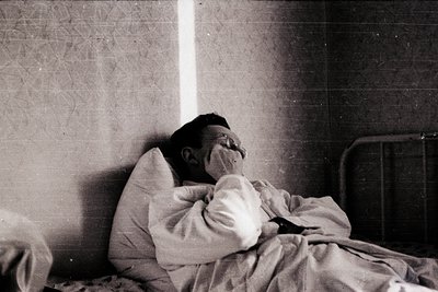 A person rests on a simple metal-framed bed in a minimalist room, wrapped in a blanket. The tiled wall and worn linoleum floo...
