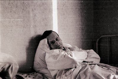 Mid-20th century hospital room interior featuring a patient reclining on a metal-framed bed with a white blanket. Plain tiled...