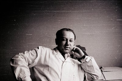 Portrait of a man in a white lab coat leaning on a metal railing, mid-conversation on a phone. Mid-20th century industrial or...