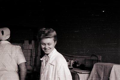 Vintage black-and-white photo of two women in indoor setting, likely a kitchen or workshop. Woman in foreground wears a short...