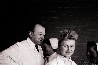 Black-and-white portrait of two individuals in formal attire, likely 1950s–1960s. Man in white lab coat, woman in high-necked...