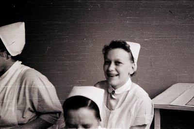 Black-and-white vintage photo of two nurses in classic caps and collared uniforms, likely mid-20th century. One nurse smiles ...