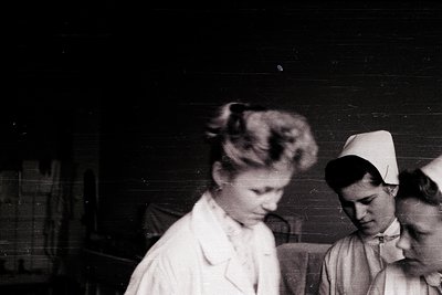 Three nurses in early 20th-century uniforms—white caps, collared gowns—pose indoors, likely a hospital or clinic. Blurred bac...