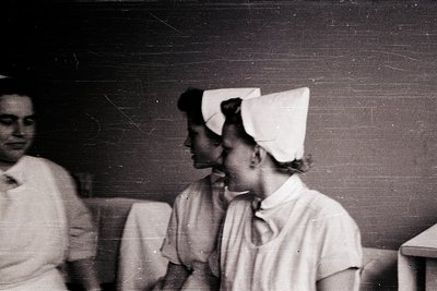 Two nurses in vintage white uniforms and caps pose indoors, likely mid-20th century. Uniforms feature high-necked collars and...