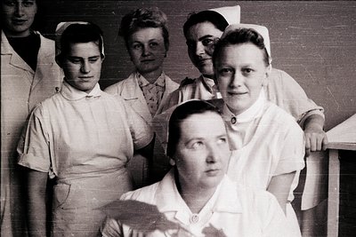 Five nurses in early 20th-century uniforms pose indoors, likely a clinic or hospital. White starched collars, high-necked blo...