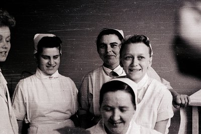 Five healthcare workers pose indoors, likely mid-20th century (1940s–1960s). Uniforms feature starched white caps and collare...