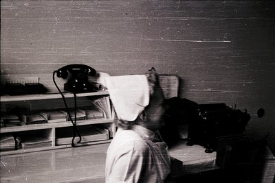 Vintage black-and-white office scene featuring a nurse in uniform attending to a vintage rotary phone on a rolling cart. Shel...