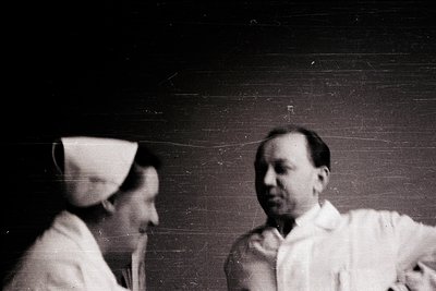 Vintage black-and-white portrait of two individuals in medical attire—nurse’s cap and lab coat—suggesting a clinical or hospi...