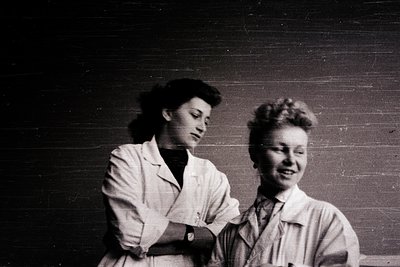 Two women in 1940s-style lab coats pose indoors against a plain wall. Both wear short, voluminous hairstyles typical of mid-c...