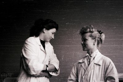 Two women in 1940s-style lab coats pose indoors, leaning toward each other in a candid moment. The woman on the left wears he...