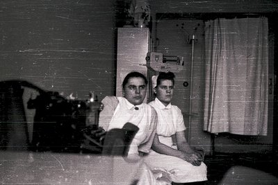 Vintage black-and-white photo of two nurses in mid-20th century scrubs, seated beside a vintage medical cabinet and sewing ma...